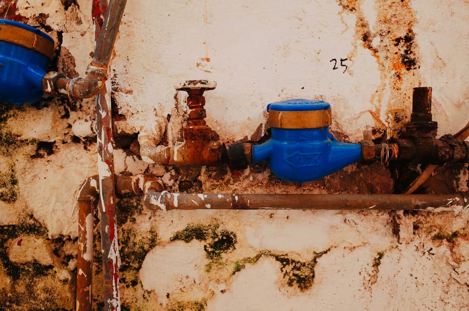 A corroded gate valve and a water meter on rusted steel pipe along a stained wall