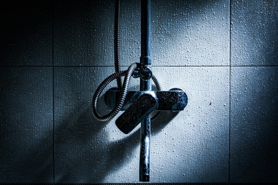 A wall-mounted shower mixer and hose on a tiled wall in cold blue light